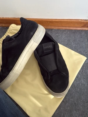 Svarta slip-on sneakers från Axel Arigato - Svarta slip-on sneakers från Axel Arigato i mocka med vit tjock sula. Minimalistisk design utan snörning och diskret logga på plösen. Perfekta för en clean och stilren look. Gått sönder lite på sulan tyvärr. Storlek 41 men passar 42 också då det är läder där i
