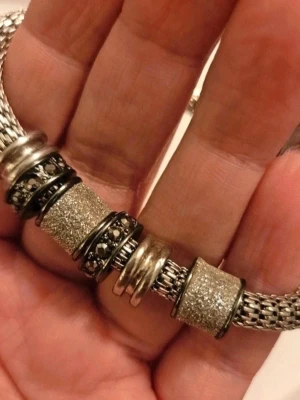 Silvrig chunky armband med glitterdetaljer - Säljer ett chunky armband i silvrig metall med flera glittriga och mönstrade berlocker. Armbandet har en flätad kedja och stora, blanka och glittriga detaljer som ger en cool och modern look. Perfekt accessoar för att lyfta din outfit.  Kan även hämtas I Partille  J