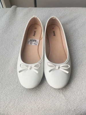 Vita ballerinaskor med rosett - Snygga vita ballerinaskor i storlek 37 med rund tå och söt rosett framtill. HELT NYA endast använda 1 gång!!!!