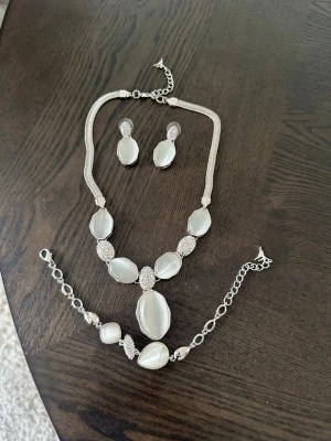 Set med silverfärgade smycken - Elegant set med halsband, armband och örhängen i silverfärg med stora vita stenar och gnistrande detaljer. Perfekt för att ge en lyxig touch till din outfit. Materialet är metall och syntetiska stenar, designen är modern och stilren.