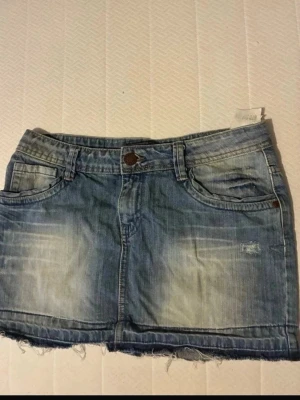 Blå jeanskjol med rå kant - Cool blå jeanskjol från H&M med slitna detaljer och rå, fransig kant längst ner. Kjolen har klassiska fickor fram och bak samt knapp och dragkedja i midjan. Perfekt för dig som gillar en avslappnad och trendig look.