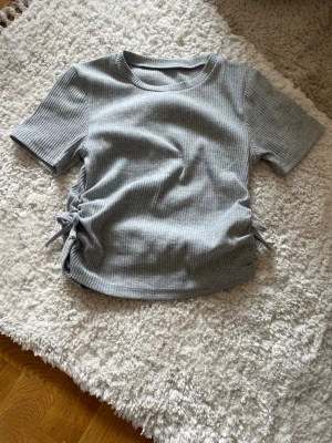 Grå ribbad topp med snörning - Säljer en grå ribbad topp med korta ärmar och snörningsdetaljer på båda sidor. Toppen har en rund halsringning och är tillverkad i ett mjukt, stretchigt material som ger en slim passform. För liten för mig, men jätte skön