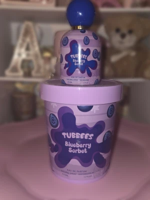 Tubbees Blueberry Sorbet parfym - Tubbees Blueberry Sorbet är en parfym med en söt och fruktig doft av blåbär. Flaskan är lila med blå detaljer och har ett lekfullt designmönster med blåbär och splash-effekter. Parfymen kommer i en rund flaska med blått klotformat lock och en matchande lila förpackning.