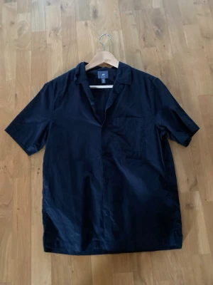 Svart kortärmad linneskjorta skjorta från H&M - Svart skjorta från H&M med regular fit och korta ärmar. Skjortan har klassisk krage, bröstficka och knäppning framtill. Perfekt för en clean och stilren look. Linne