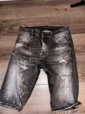 Grå s jeansshorts från DSQ2 - Snygga grå jeansshorts från DSQ2 med slitningar, färgstänk och coolt tvättad look. Klassisk femficksmodell med knappgylf och råa benslut. Perfekta för dig som gillar streetstyle och vill sticka ut lite extra.
