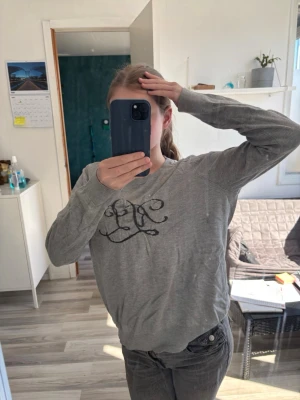 Grå sweatshirt med svart tryck - Grå sweatshirt med rund hals och svart tryck framtill i form av ett snirkligt mönster. Tröjan har långa ärmar och en avslappnad passform, perfekt för en chill och stilren look.