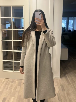 Grå lång kappa i ullmix - Säljer en stilren och lång beige kappa med rak passform. Jackan har klassisk krage och är helfodrad, perfekt för kyligare dagar. Materialet är mjukt och känns lyxigt, troligtvis en ullblandning. Passar dig som gillar en clean och tidlös look.
