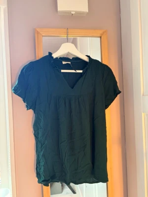 Mörkgrön blus med volangkrage - En mörkgrön blus med korta ärmar och v-ringning. Blusen har en söt volangdetalj vid kragen och ett lätt, luftigt tyg som ger en avslappnad känsla. Perfekt att matcha med jeans eller kjol för en trendig look.