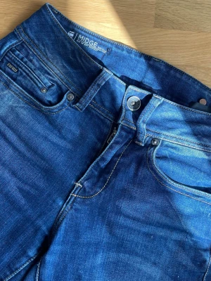Blå G-Star RAW Midge jeans - Säljer ett par snygga blå jeans från G-Star RAW, modell Midge. Jeansen har klassisk femficksdesign, silverfärgad knapp och dragkedja samt diskreta slitningar för en cool look. De här har då blivit förkorta för mig vilket är jättesynd för jag älskade de här jeansen.