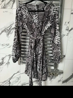 Kort omlottklänning med ormmönster - Säljer en kort omlottklänning med svart, grått och vitt ormmönster. Klänningen har långa ärmar, v-ringning och ett knytband i midjan för snygg passform. Inga skador eller defekter. 