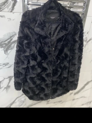 Svart fuskpäls kappa från Vero Moda - Snygg svart kappa i fuskpäls från Vero Moda. Kappan har ett vågigt mönster i pälsen, klassisk krage och knäppning framtill. Inga skador eller defekter. 