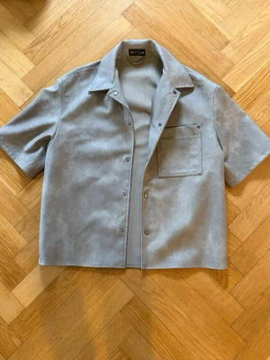 Grå kortärmad skjorta i mocka-look - Stilren grå skjorta från zara i mocka-liknande material. Skjortan har korta ärmar, tryckknappar framtill och en bröstficka. Perfekt för dig som gillar minimalistisk och modern stil.