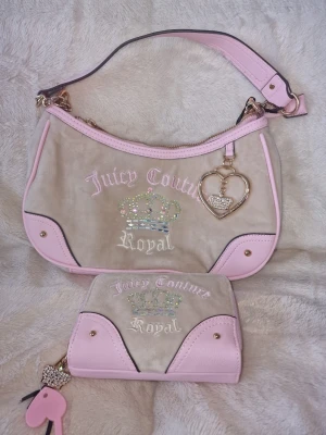 Juicy Couture beige och rosa handväska - Juicy Couture handväska i beige sammet med ljusrosa detaljer i skinnimitation. Väskan har broderad krona med strass, texten 'Juicy Couture Royal' och ett hjärtformat hänge. Matchande plånbok medföljer, även den med broderi och rosa detaljer.