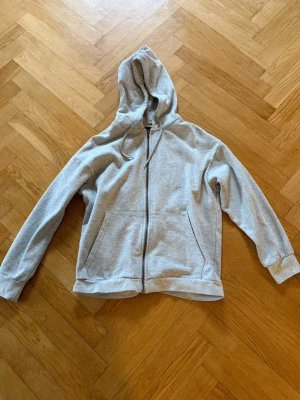 Ljusgrå zip-hoodie med huva - En stilren ljusgrå hoodie med dragkedja framtill och två fickor. Klassisk modell med huva och dragsko, samt ribbade muddar vid ärmslut och nederkant. Perfekt för en avslappnad och trendig look.