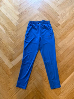 Blå träningsbyxor från Nike - Snygga blå träningsbyxor från Nike med svart logga på vänster lår. Byxorna har resår i midjan och är tillverkade i ett lätt och stretchigt material som passar perfekt för sport och träning.