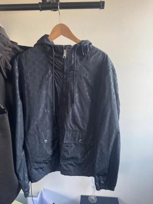 Svart vindjacka från Gucci - Svart vindjacka från Gucci med diskret GG-mönster över hela tyget. Jackan har huva med dragsko, två stora fickor framtill och elastiska muddar vid ärmslut och nederkant. Perfekt för en stilren och lyxig streetwear-look.