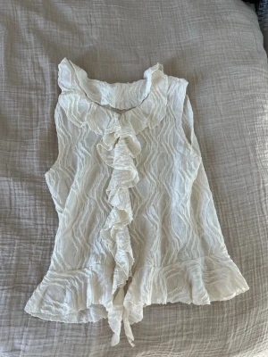 Vit volangblus från Gina Tricot - Supersöt vit blus från Gina Tricot med volanger längs krage och framtill. Blusen är ärmlös och har ett lätt transparent, strukturerat tyg som ger en romantisk känsla. Perfekt att styla med jeans eller kjol för en trendig look.