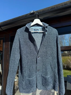 Stickad Kavaj - Mysig grå/blå stickad kavaj med två knappar framtill och två framfickor. Kavajen har en avslappnad passform, sjalkrage och är tillverkad i grovstickat material. Perfekt för en chill och stilren look. Säljer då den är lite för liten för mig vid axlar och armar.