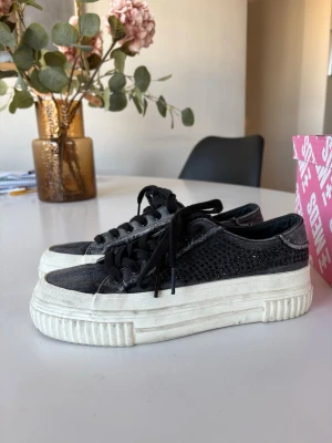 Svarta platåsneakers i jeans med stenar - Säljer ett par svarta sneakers i jeans från Zara med platåsula och glittriga detaljer på sidorna. Skorna har svarta skosnören och en vit, kraftig sula. Perfekta för dig som vill sticka ut med en cool och trendig look. I stortsätt aldrig använda.