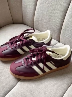 Adidas Samba vinröda sneakers - Snygga Adidas Samba sneakers i vinrött läder med klassiska vita ränder på sidorna och vit häl. Skorna har låg profil, gummisula och retrovibbar. Perfekta för dig som gillar streetstyle och vill sticka ut med en unik färg på dina sneakers.