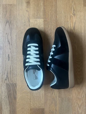 Svarta sneakers från Maison Margiela - Maison Margiela sneakers i svart läder och mocka med vita skosnören och klassisk gummisula i brunt. Låg modell med retrodesign och detaljerad yttersula för extra grepp. Perfekt för dig som gillar stilrena och tidlösa sneakers.