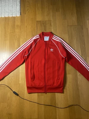 Röd Adidas track jacka - Snygg röd Adidas track jacket med klassiska vita ränder längs ärmarna och vit Adidas-logga på bröstet. Dragkedja framtill och ribbade muddar. Perfekt för en sportig och retro look.
