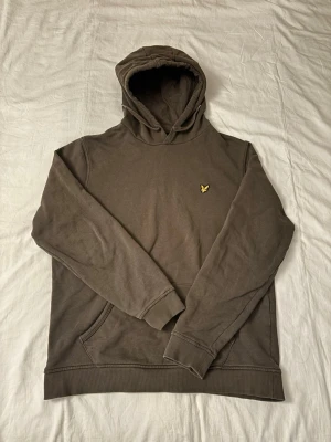 Olivgrön hoodie från Lyle & Scott - Snygg olivgrön/khaki hoodie från Lyle & Scott med klassisk gul logga på bröstet. Hoodien har huva, känguruficka och ribbade muddar vid ärmslut och nederkant. Perfekt för en avslappnad och stilren look. Är storlek XL men upplevs som L
