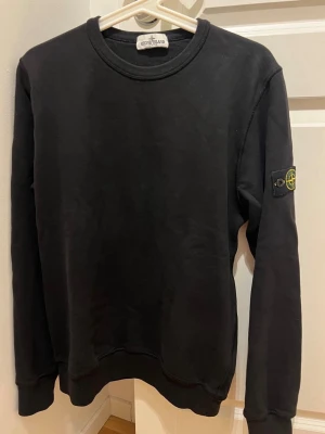 Svart sweatshirt från Stone Island (JR) - Svart sweatshirt från Stone Island med klassisk rund hals och ribbade muddar. Ikonisk logotyp-patch på vänster ärm. Perfekt för en stilren och avslappnad look. Juniorstorlek, 170, men stor i storleken och passar även en S. Enbart använd ett fåtal gånger. Katt finns i hemmet. Digital orderbekräftelse kan lämnas vid önskan.