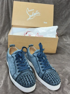 Blåa Christian Louboutin sneakers - Snygga blå sneakers från Christian Louboutin med coola nitar på tån och matchande blå skosnören. Ovandelen är i mockaliknande material och sulan är vit. Perfekta för dig som vill sticka ut med en lyxig och edgy stil.