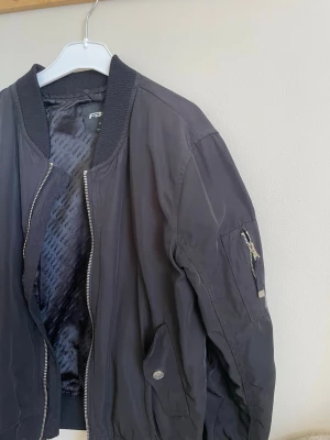 Svart bomberjacka från FB Sister - Stilren svart bomberjacka från F&F med klassisk ribbad krage, muddar och dragkedja framtill. Jackan har fickor med tryckknapp och en ärmficka med dragkedja. Materialet är glansigt och lätt, perfekt för en cool streetstil.
