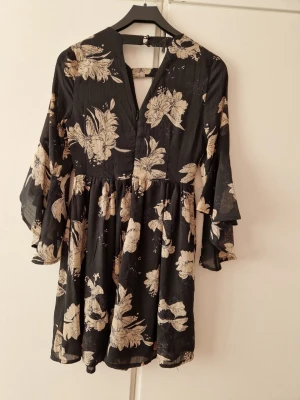 Svart blommig klänning med volangärm - Svart klänning med beige blommönster och v-ringning. Klänningen har volangärmar och en lätt utställd kjol. Tillverkad i ett tunt, flowy material som ger en luftig känsla. Perfekt för dig som gillar romantiska och trendiga plagg.