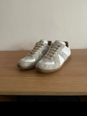 Maison margielas gats - Snygga sneakers från Maison Margiela i vitt läder och beige mocka med klassisk gummisula. Modellen har låga skaft, neutrala färger och stilren design som passar till det mesta. Perfekta för dig som gillar minimalistisk och tidlös stil.