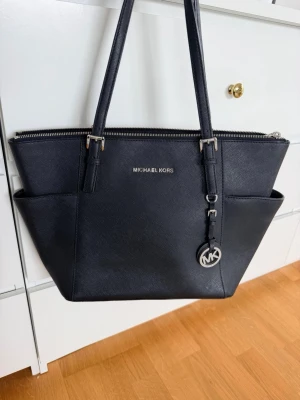 Svart handväska från Michael Kors - Stilren svart handväska från Michael Kors i strukturerat läder med silverfärgade detaljer och logotyp framtill. Väskan har två långa axelband, dragkedja upptill och praktiska sidofack.