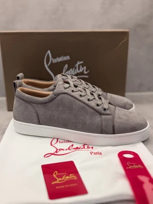 Gråa sneakers från Christian Louboutin - Stilrena gråa sneakers från Christian Louboutin i mocka med vita sulor och gråa skosnören. Klassisk låg modell med diskreta sömmar och lyxig känsla. Perfekta för dig som vill ha en clean och exklusiv look.
