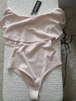 Ljusrosa off shoulder body Boohoo - Trendig ljusrosa body från Boohoo med off shoulder-design och volangdetalj upptill. Tillverkad i mjukt, stretchigt material som sitter snyggt på kroppen. Perfekt att styla med jeans eller kjol för en cool look.
