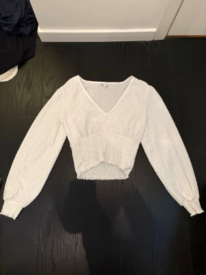 Vit smockad blus med v-ringning - Trendig vit blus med smockad midja och ärmslut, v-ringad hals och lång ärm. Blusen har en lätt strukturerad yta och en snygg, något kortare passform. Perfekt att styla med jeans eller kjol för en fräsch look.
