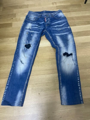 Blå slitna jeans från Dsquared2 - Riktigt snygga blå jeans från Dsquared2 med slitningar, hål på båda knäna och coola färgstänk. Jeansen har rak passform och klassisk femficksdesign. Perfekta för dig som gillar en edgy och trendig streetstil.