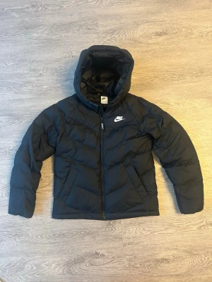Svart dunjacka med huva från Nike - Snygg svart dunjacka från Nike med huva och dragkedja framtill. Jackan har en klassisk puffer-design och Nike-logga på bröstet. Passar perfekt för kalla vinterdagar och har en sportig look som är enkel att matcha. Storlek L för tonåringar och 147-158cm 