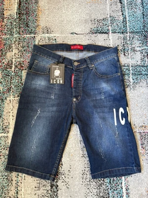 Blå jeansshorts från Dsquared2 ICON - Snygga blå jeansshorts från Dsquared2 ICON-kollektionen med slitna detaljer och vit text på ena benet. Klassisk femficksmodell med knappgylf och röd Dsquared2-tag vid fickan. Perfekta för en trendig streetstyle-look.