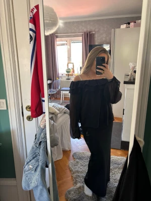 Svart blus - Elegant svart blus med offshoulder-modell och volangdetaljer längs överdelen. Blusen har en rak och flowy passform och är tillverkad i ett tunt, lätt material som ger en snygg siluett.