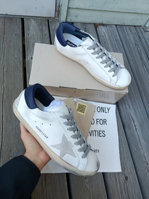 Golden Goose vita sneakers med blå detalj - Golden Goose sneakers i vitt läder med grå stjärna på sidan, grå skosnören och blå hälkappa. Klassisk låg modell med slitna detaljer och gummisula. Märket syns på sidan och hälen. Perfekta för dig som gillar en avslappnad men trendig stil.