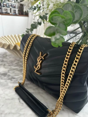 Svart trendig axelväska med guldkedja design Saint laurent - Snygg svart axelväska i quiltat läder med YSL-logga i guld framtill. Väskan har en elegant guldfärgad kedjerem med läderdetalj för axeln. Perfekt för att lyfta din outfit med en lyxig touch. Lulu modell.