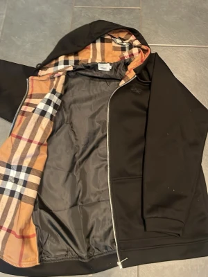 Svart hoodie med Burberry-mönstrad huva - Snygg svart hoodie med dragkedja från Burberry. Utsidan är helsvart medan insidan av huvan har det klassiska Burberry-rutiga mönstret i beige, svart och rött. Hoodien har fickor framtill. Det är storlek Xl och helt oanvänd. Pris kan diskuteras.