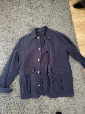 Mörkblå overshirt med fickor - Stilren mörkblå overshirt med klassisk krage och stora fickor framtill. Jackan har knäppning med ljusa knappar och raka ärmar. Perfekt för lager-på-lager och enkel att matcha med olika outfits.