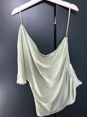 Ljusgrön asymmetrisk linnetopp - Trendig ljusgrön linnetopp med en axelbandslös design. Toppens material är mjukt och har ett snyggt fall. Perfekt för dig som gillar unika och stilrena plagg med en twist.