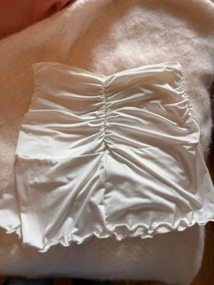 Vit bandeau tubtopp med rynk - Säljer en vit bandeau tubtopp med snygg rynkdetalj framtill och vågig kant nertill. Toppen är axelbandslös och har en mjuk, stretchig känsla som sitter bekvämt. Perfekt att styla med jeans eller kjol för en trendig look. (Har en smink fläck men går bort i tvätten! ) 