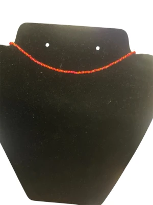 Rött pärlhalsband med små pärlor - Ett tunt halsband med små röda och orange pärlor i glas. Halsbandet har ett enkelt och stilrent utseende med ett guldfärgat spänne. Perfekt för att addera lite färg till din outfit.