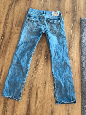 Blå jeans från True Religion - Säljer ett par klassiska blå jeans från True Religion med raka ben och snygga detaljerade bakfickor. Jeansen har ljus tvätt och synliga sömmar, samt logga på bakfickan och patch i midjan. Perfekta för en avslappnad och trendig look. Pris kan diskuteras!