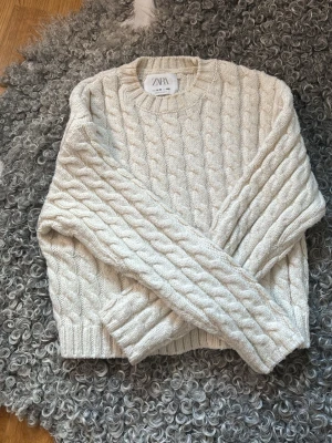 Vit kabelstickad tröja från Zara - Kabelstickad tröja från Zara i mjuk, vit ullblandning. Tröjan har rund halsringning, ribbade muddar och ett klassiskt flätmönster över hela plagget. Perfekt för dig som gillar en clean och cozy stil.