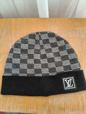 Louwis wuitton svartgrå rutmönstrad mössa - Snygg svart och grå mössa från Louwis wuitton med klassiskt rutmönster och vikt kant. L v-logga framtill på en svart patch. Stickad i mjukt material som håller dig varm och stilsäker under kalla dagar. Perfekt accessoar för dig som gillar exklusiv streetwear.